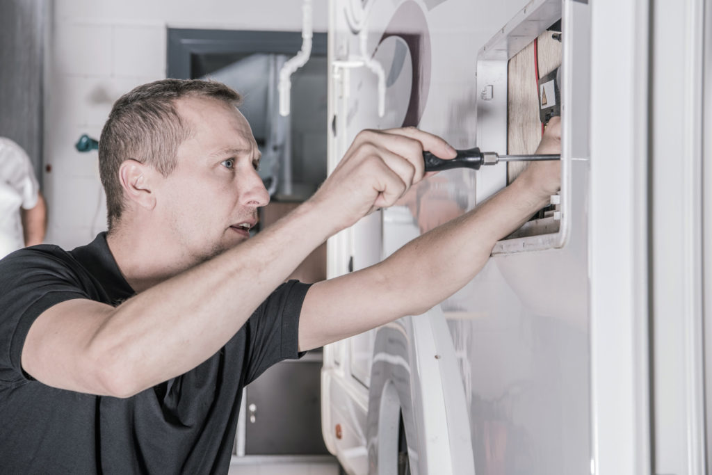 Camper Van RV Refrigerator Repair. Motorhome Appliances Maintenance. Caucasian RV Technician with Screwdriver Looking For Potential Issues Inside Outside Camper Compartment. Automotive and Travel Industry Theme.
